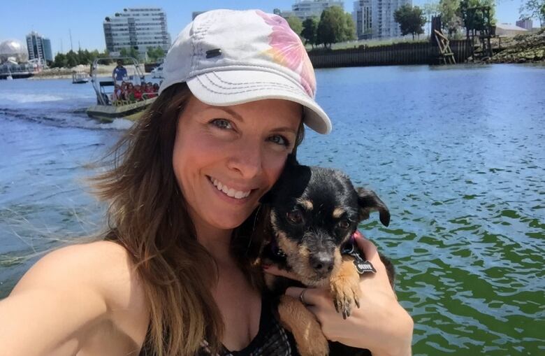 A selfie photo shows a woman with long brown hair under a white baseball cap holding a tiny black and brown dog. She is wearing a black tank top and positioned in front of False Creek in Vancouver, with the condo towers of Olympic Village in the background.