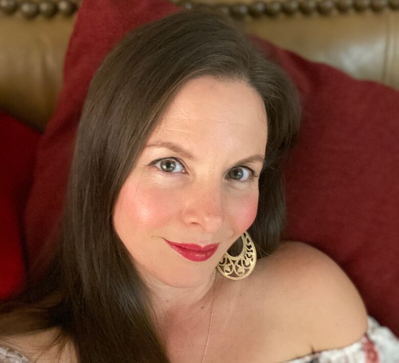 A smiling woman with long brown hair is shown in close-up. She is wearing bright red lipstick, gold earrings, and an off-the-shoulder peasant style top.