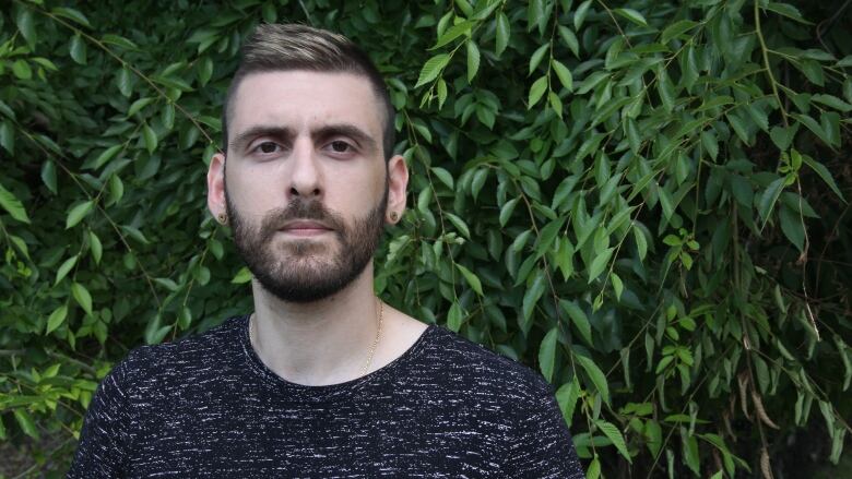 A bearded man stands in front of a leafy green tree, looking toward the camera.