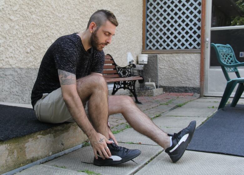 A man wearing a T-shirt and shorts sits outside a house, lacing up his running shoes.