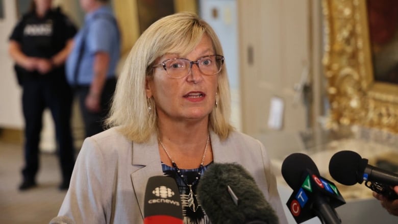 A woman wearing glasses stands with microphones in front of her.