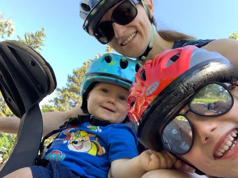 A woman poses with a toddler and an older child. All three are wearing helmets.