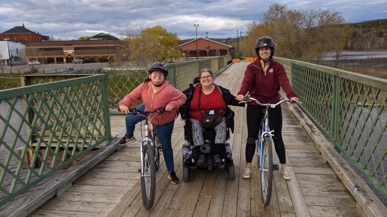 A young child on a bicycle, a teenager on a bicycle, and a woman in a wheelchair are pictured outside on a boardwalk.