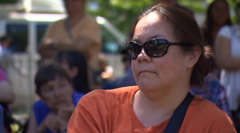 A woman wearing an orange t shirt and sunglasses stares off into the distance.