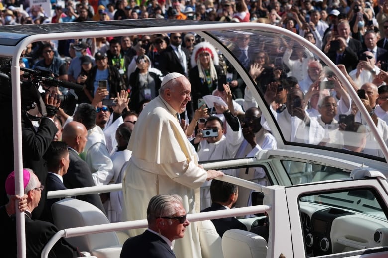 Man in white robes rides in open air vehicle through crowds