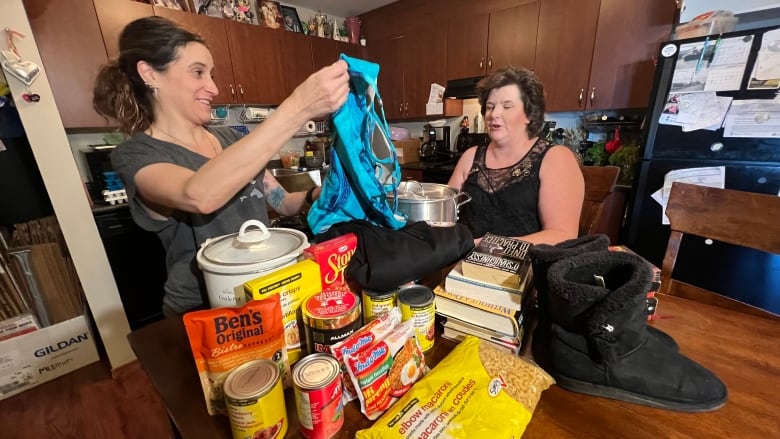 Two women stand in a kitchen behind a table full of food, household items and clothing. One woman with a ponytail holds up a shirt.