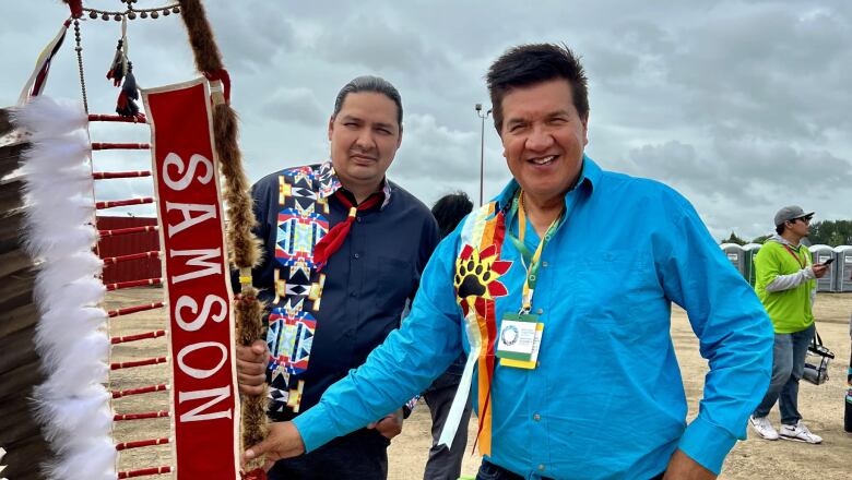 Two Indigenous man are smiling and holding a banner on a stick with feathers that reads 