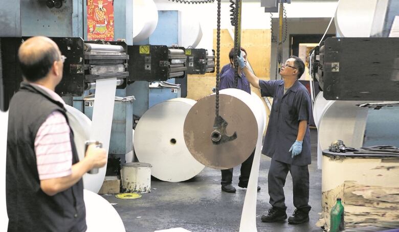 A group of men stare at rolls of paper at a printing press.