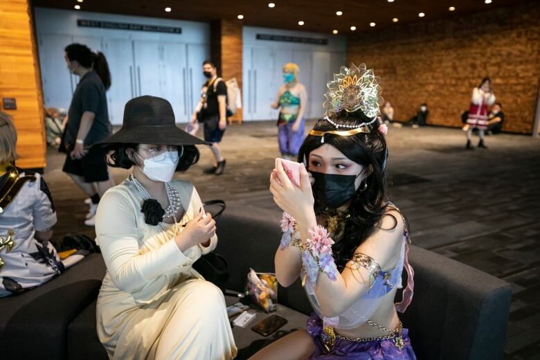 Two women wearing cosplay help each other with makeup, with other people in the background waiting in line.