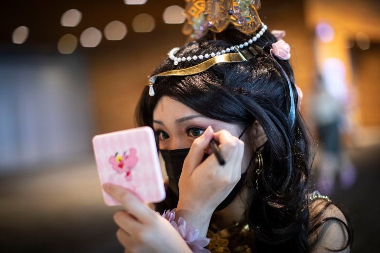 A woman applies an eyelash pencil to herself, with a Pink Panther mirror. She is dressed up like an anime character and has a crown on.