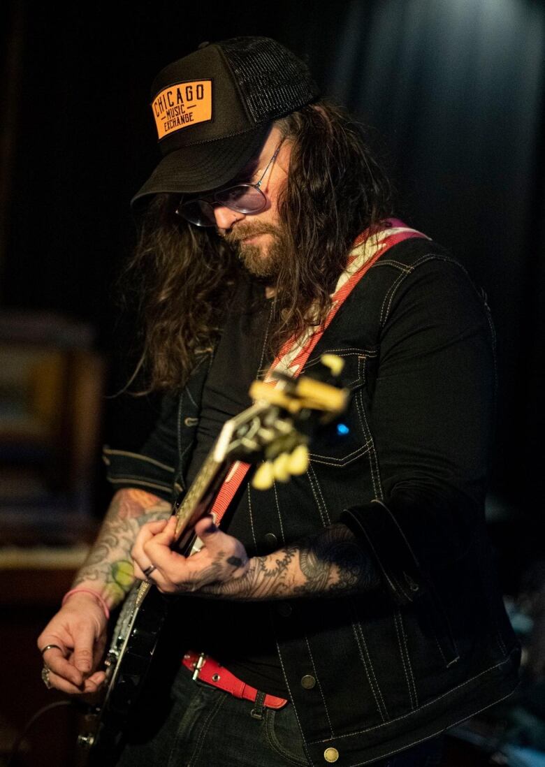 A long-haired man in dark clothes, wearing glasses and a baseball cap, plays electric guitar.