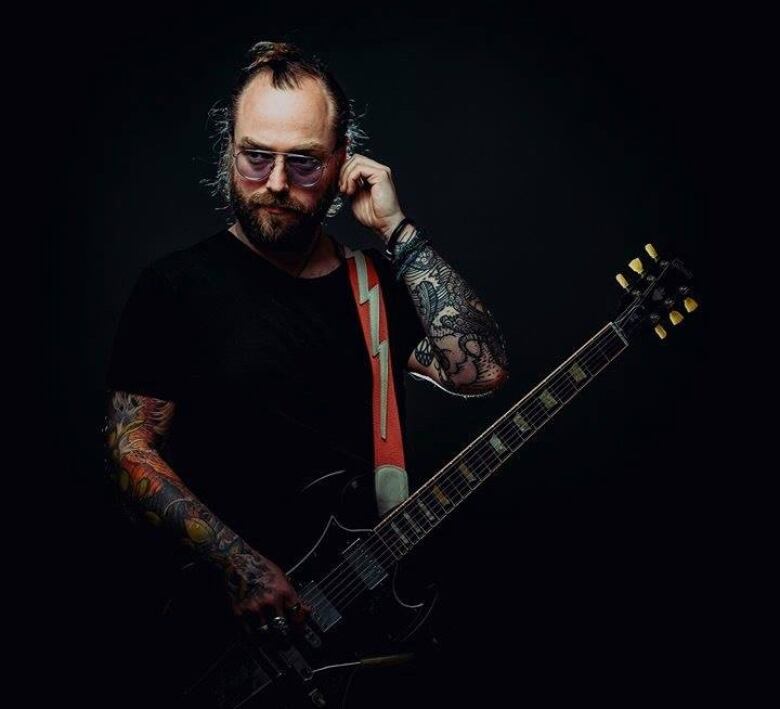 A bearded man wearing glasses and a black T-shirt holds an electric guitar.