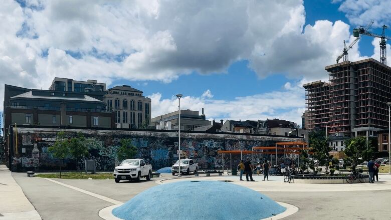 A park on a sunny day, with two white trucks, people standing underneath an orange overhang, with a long, graffiti-ed building in the background. 