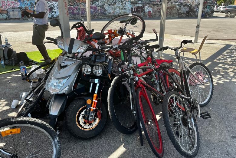 A pile of bicycles and electric scooters in the shade of a splash park overhang. 