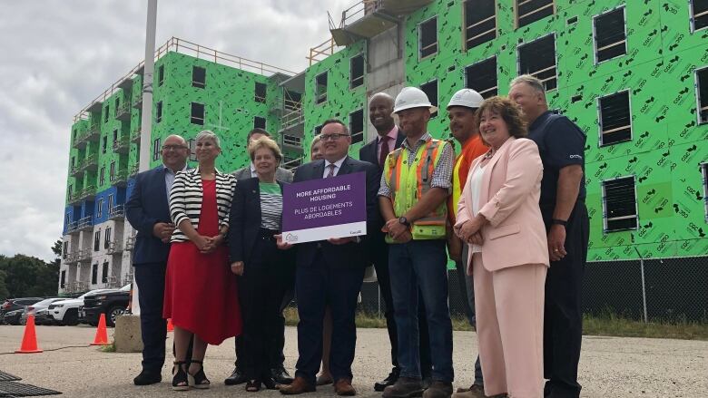 Group of politicians pose for a photo in front of an apartment building under construction. MP Bryan May in the middle front of the group holds a sign that reads 'more affordable housing.'