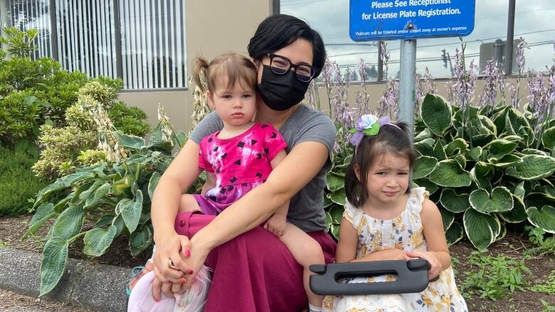 A woman in a mask sits on a pavement with two children.