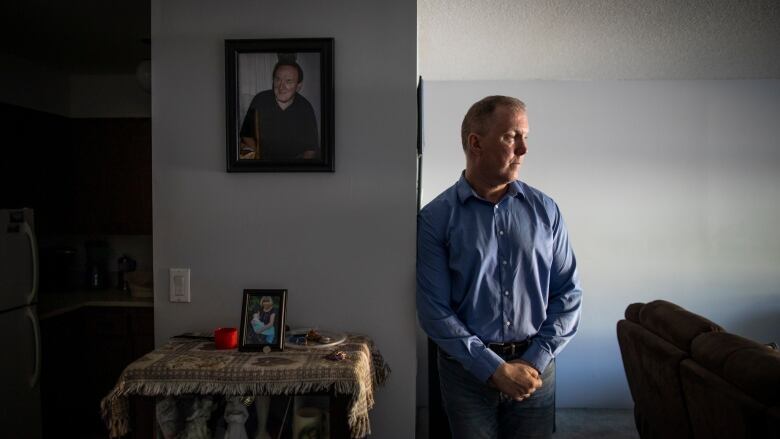 Norm leans against a wall. He is wearing a blue long-sleeved shirt.