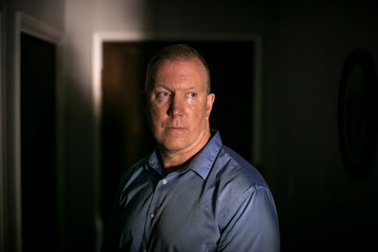 Norm looks away from the camera in a dimly-lit room. He is wearing a blue long-sleeved shirt.