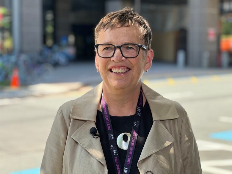 A white woman with her brown hair in a pixie cut is wearing glasses and a beige jacket over a black top