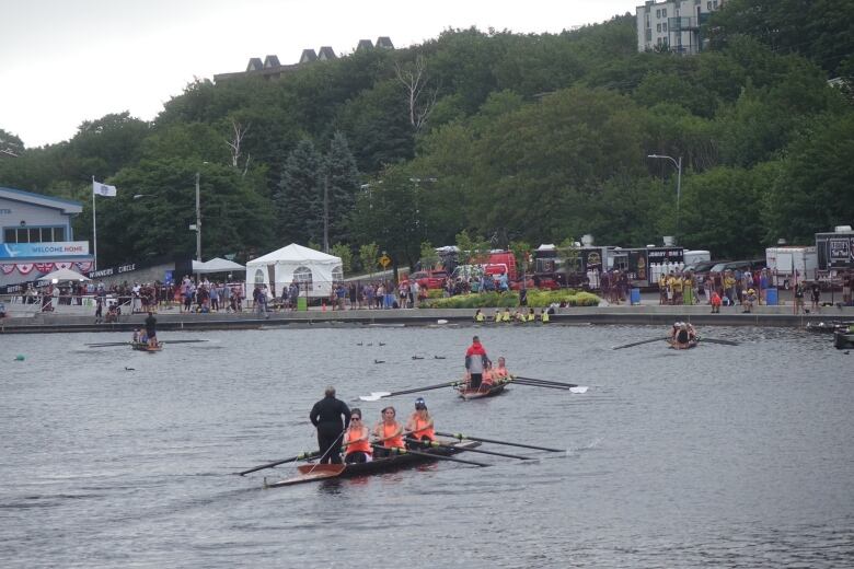 Five row boats are various distances from the shore of a lake. Food trucks, tents and spectators are on the shore.