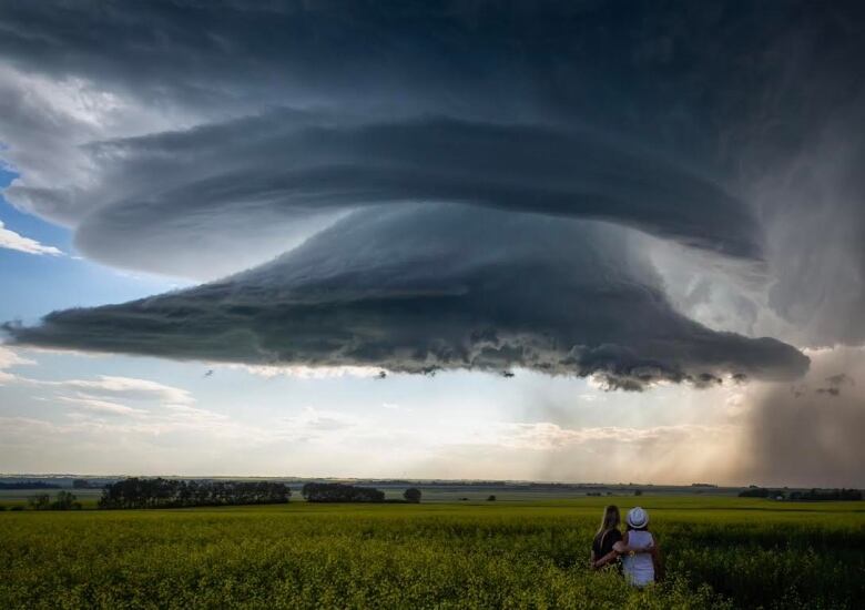 two women embrace in an empty field with dark clouds above. 