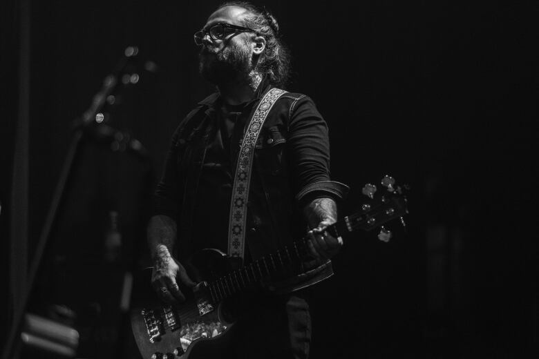 In shadowy black and white, a man in dark clothes plays an electric guitar.