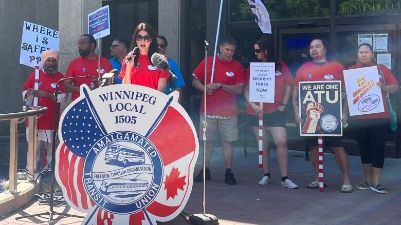 A woman with long dark hair and wearing sunglasses and a red shirt is speaking into a microphone, standing behind a podium showing the local for Amalgamated Transit Union Winnipeg Local 1505. Several people wearing the same red shirt and holding signs are standing behind her.