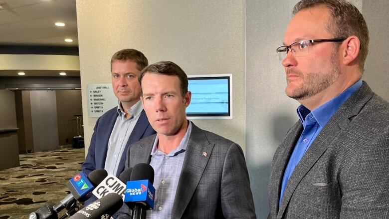 Three men in blazers and collared shirts stand in front of a group of microphones while meeting with news reporters in a Regina hotel. The first looks into the camera with his hands clasped in front of him; the second speaks into the microphones with his hands open before him; the third stands in profile with a concerned expression. A hallway is seen on the left side of the frame.