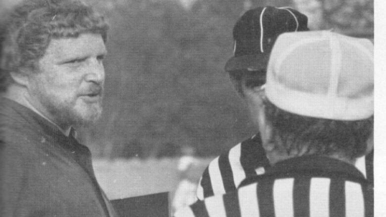 A man with curly hair holding a football and speaking with a referee.