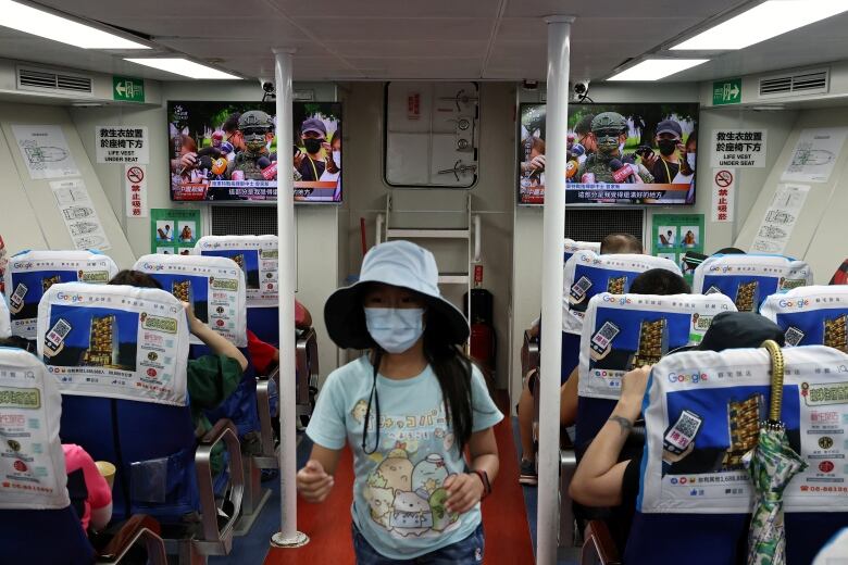 Rows of people, seated on a ferry, watch TV news coverage on a pair of screens.