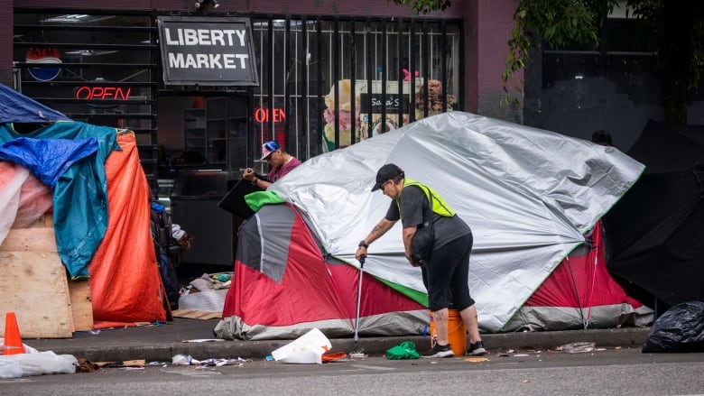 A person in a high-vis jacket removes garbage from a pavement with a tent on it, opposite a store.