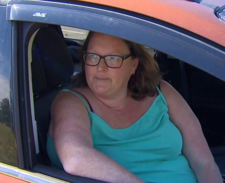 A middle-aged woman sits in the passenger seat of a car. Her right elbow is propped up on the inside of the door. She is looking out of the window which is rolled down.