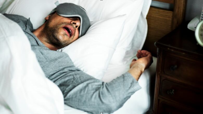 A man with dark stubble sleeps in bed with his mouth wide open. He is wearing a grey sleep mask over his eyes and grey long-sleeved pajamas.