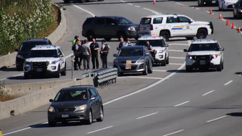 A highway is blocked off by traffic cones as police officers and vehicles are visible near parked cars.