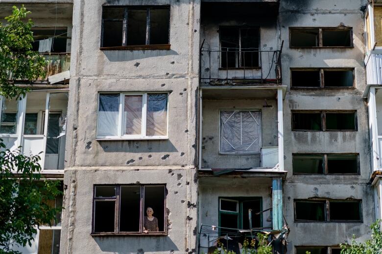 A woman stands at her window, surrounded by an apartment building filled with bullet holes.