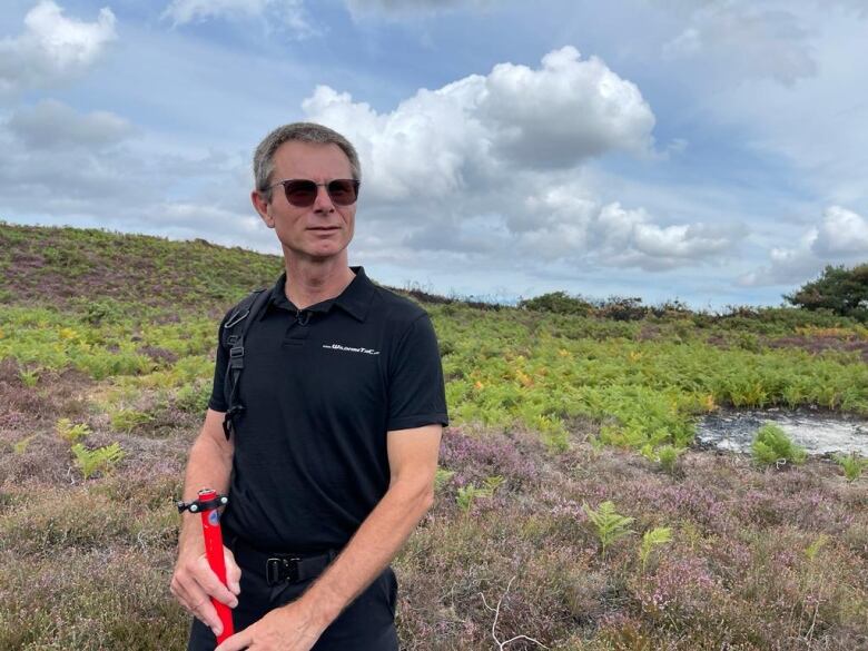 A man with short, grey hair wears sunglasses while standing in a patch of dry vegetation. 