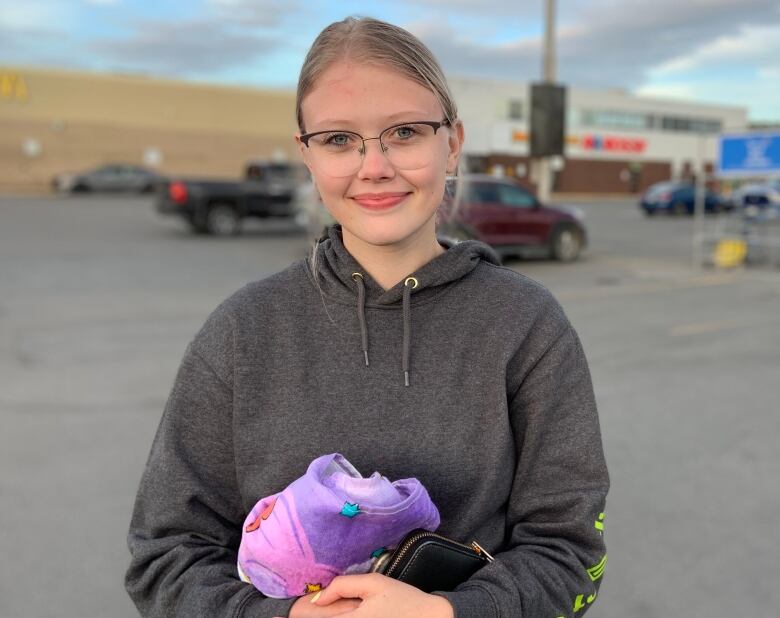 A person wearing glasses and a hoodie stands in a parking lot. She's holding a towel and a cellphone.