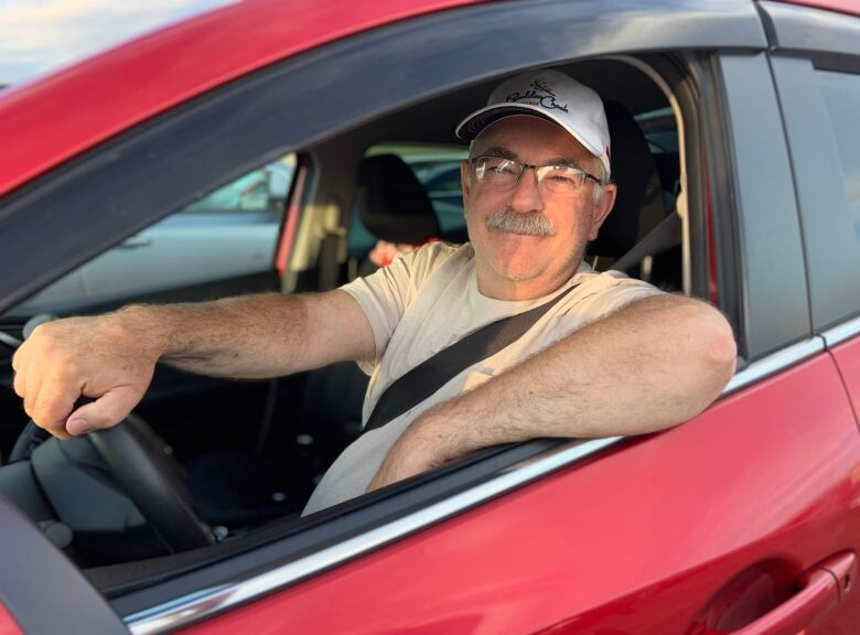 A person wearing glasses and a baseball cap sits in a red car holding the steering wheel.