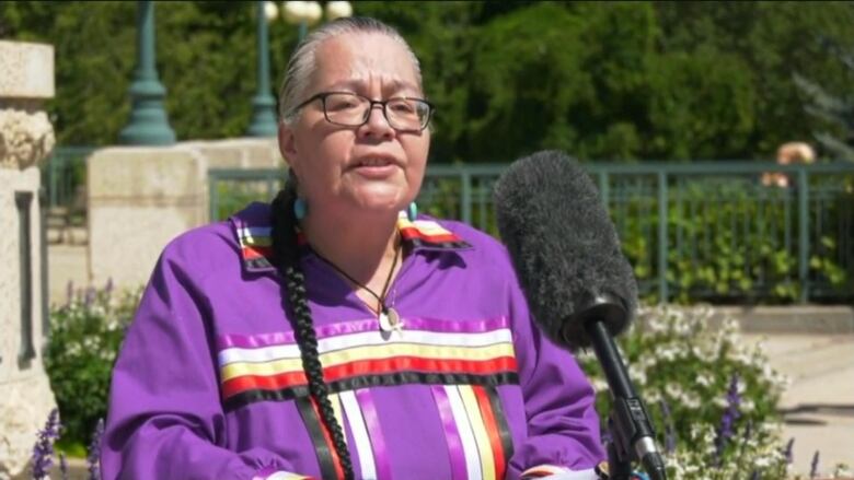 An older woman is speaking at a podium, wearing a purple shirt with ribbons across it.