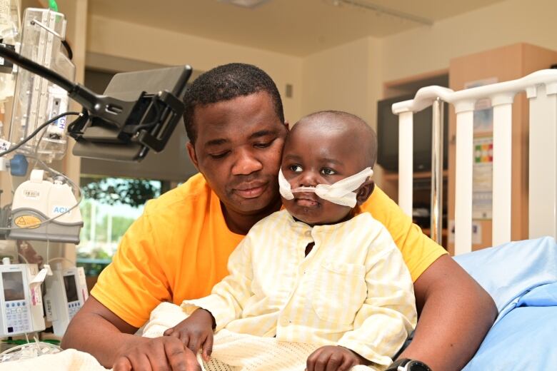 A father hugging his 2 year old son. The toddler has breathing tubers attached to his nostrils. They are in a hospital room. 