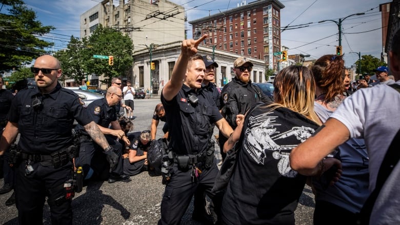 A police officer points authoritatively as others converge on residents on a sidewalk.