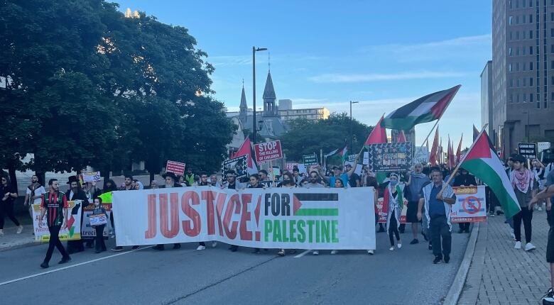 A crowd of people walk in a downtown street carrying a large sign that reads 