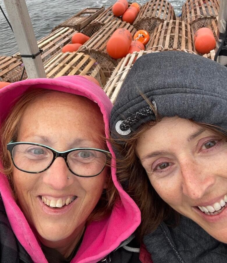 Two women smiling on a lobster boat with lots of lobster traps behind them.