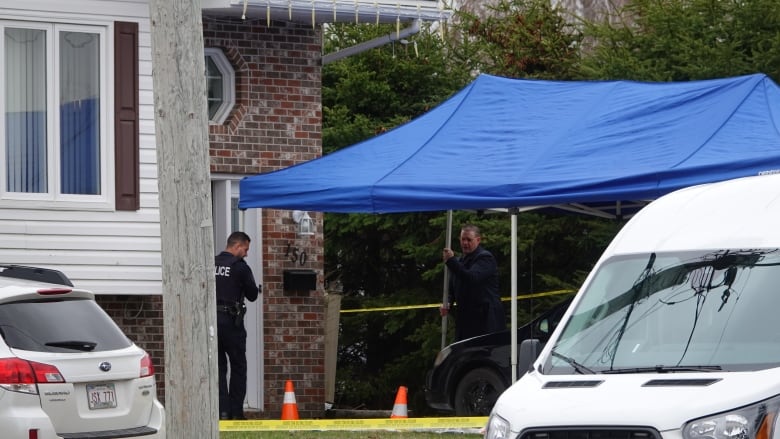 Two police officers shown outside a duplex door setting up a large blue tent. Yellow crime scene tape surrounds the area.