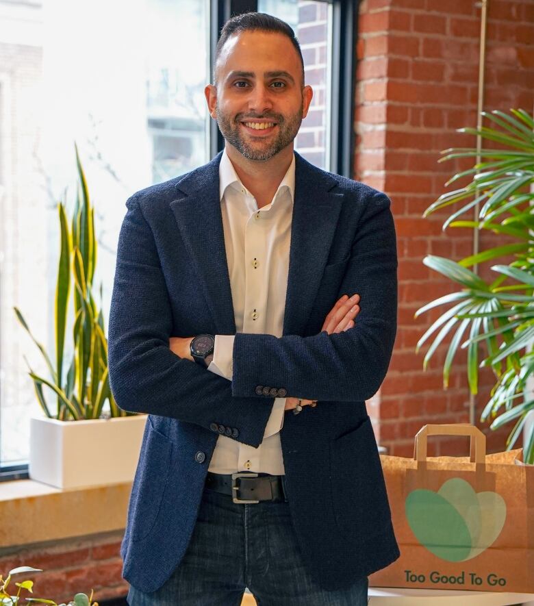 A man stands in front of a window. His arms are crossed in front of him and he has a big smile on his face. To the right of him sits a paper bag featuring the Too Good To Go food app logo.