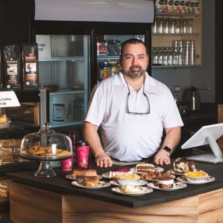 Rick Green stands inside his cafe. Glasses hang from his neck, resting on his white shirt. He's standing behind the register, surrounded by desserts and coffee