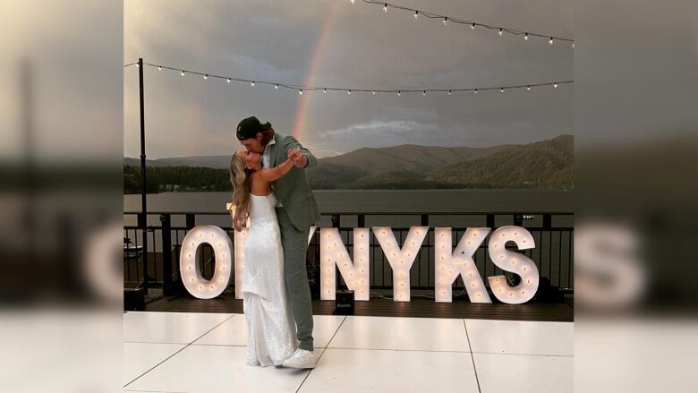 Two people kiss in front of a sign that reads 'OLYNYKS'. A rainbow is visible amid a cloudy sky.