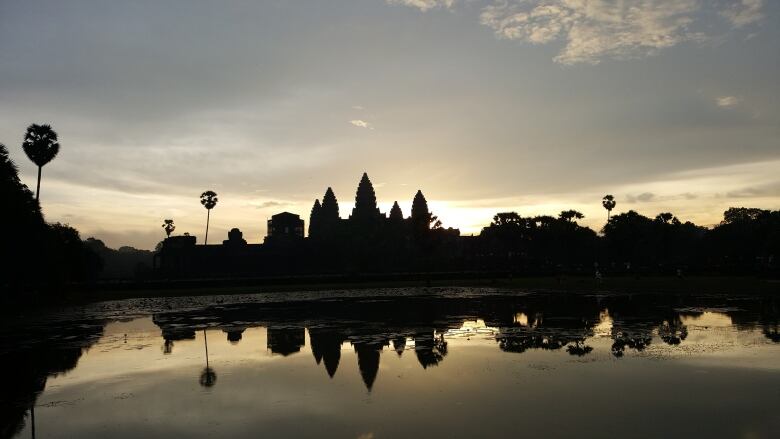 The spires of the Angkor Wat complex are visible during a sunset. The spires are in silhouette.