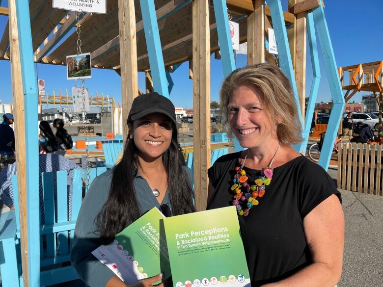 A woman with long black hair wearing a black baseball cap is smiling next to a woman with chin-length blonde hair wearing a colourful necklace and a black t-shirt. Both are holding printed copies of a report.