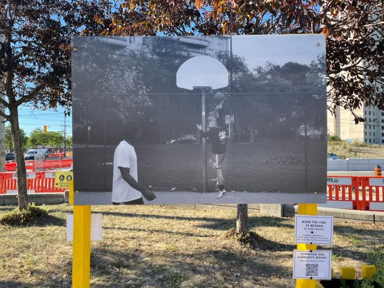 A large black-and-white photo of two children playing basketball under a hoop with no net is displayed on yellow posts. 